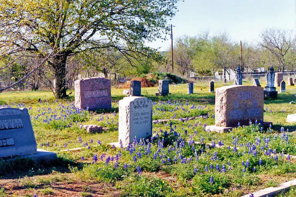 Bluebonnets in spring bloom among the gravestones of the Johnson City Masonic Cemetery, with a granite marker for Cleo Smith (1892–1949) at center and additional family stones beyond.