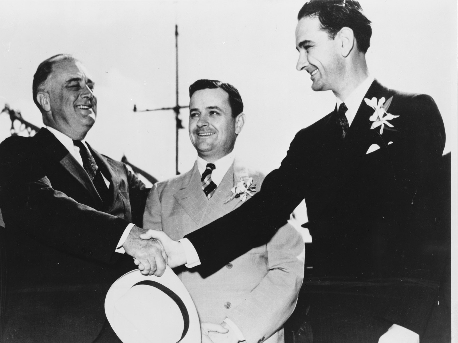 Black-and-white photograph of three men in suits outdoors. At left, President Franklin D. Roosevelt, smiling, reaches across to shake hands with a tall, dark-haired young man at right, Congressman Lyndon B. Johnson, who wears a white boutonniere and holds a light-colored hat in his other hand. Between them stands Governor James V. Allred of Texas in a light double-breasted suit, smiling at Johnson.