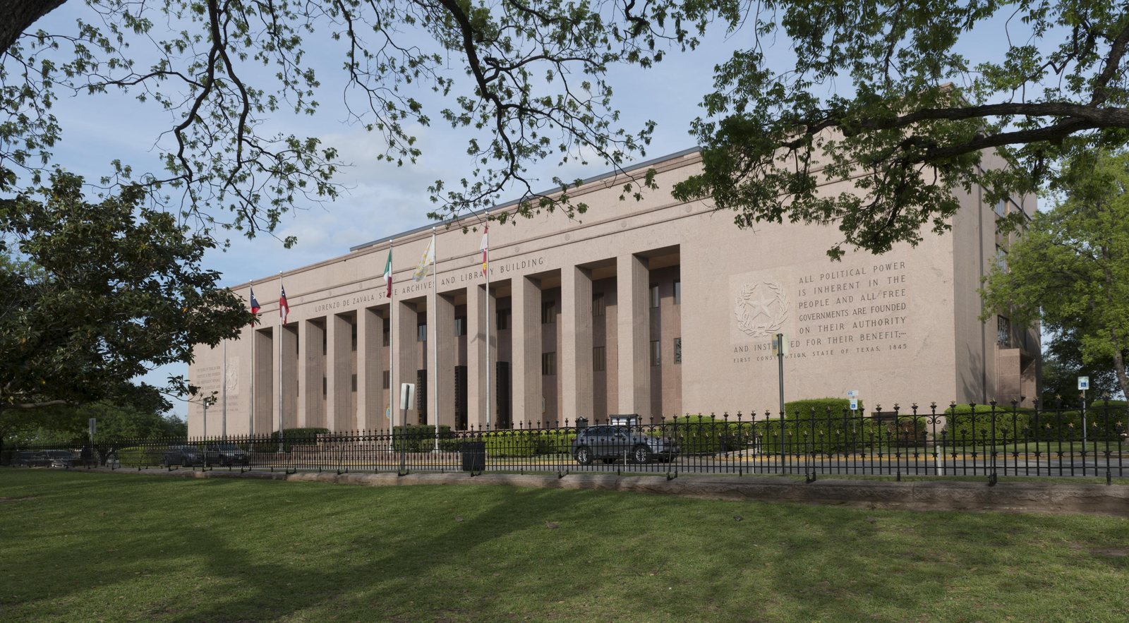 Color photograph of a long, pale pink granite building with a row of tall square columns across its front, the words LORENZO DE ZAVALA STATE ARCHIVES AND LIBRARY BUILDING cut above them, and a carved inscription beginning ALL POLITICAL POWER IS INHERENT IN THE PEOPLE on the wall at right. Flags fly on poles before it, an iron fence runs along a green lawn, and the branches of a large oak frame the top of the picture.