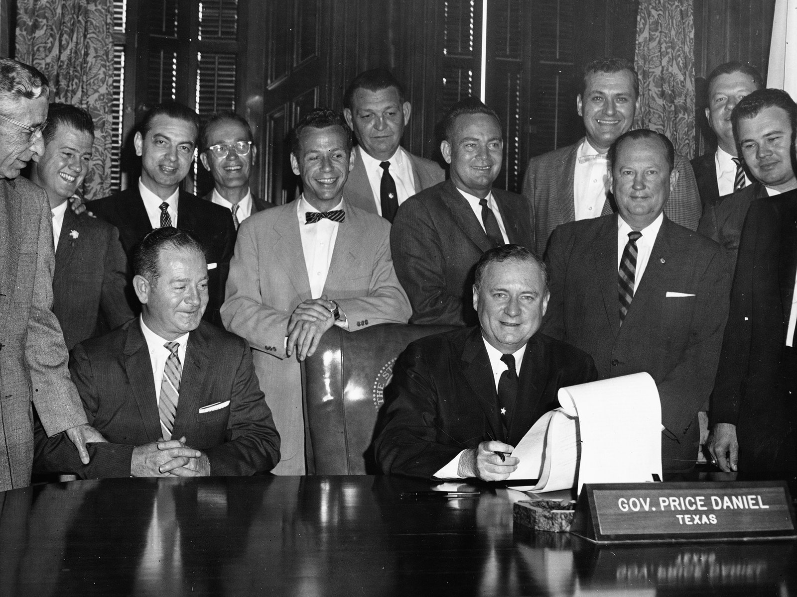 Black-and-white photograph of Governor Price Daniel seated at a polished desk in a paneled office, smiling and holding a pen over a long bill whose pages curl up beside him, with a nameplate reading GOV. PRICE DANIEL, TEXAS in front of him. Another seated man sits at his right, and more than a dozen men in suits and ties stand close behind them, most of them smiling.