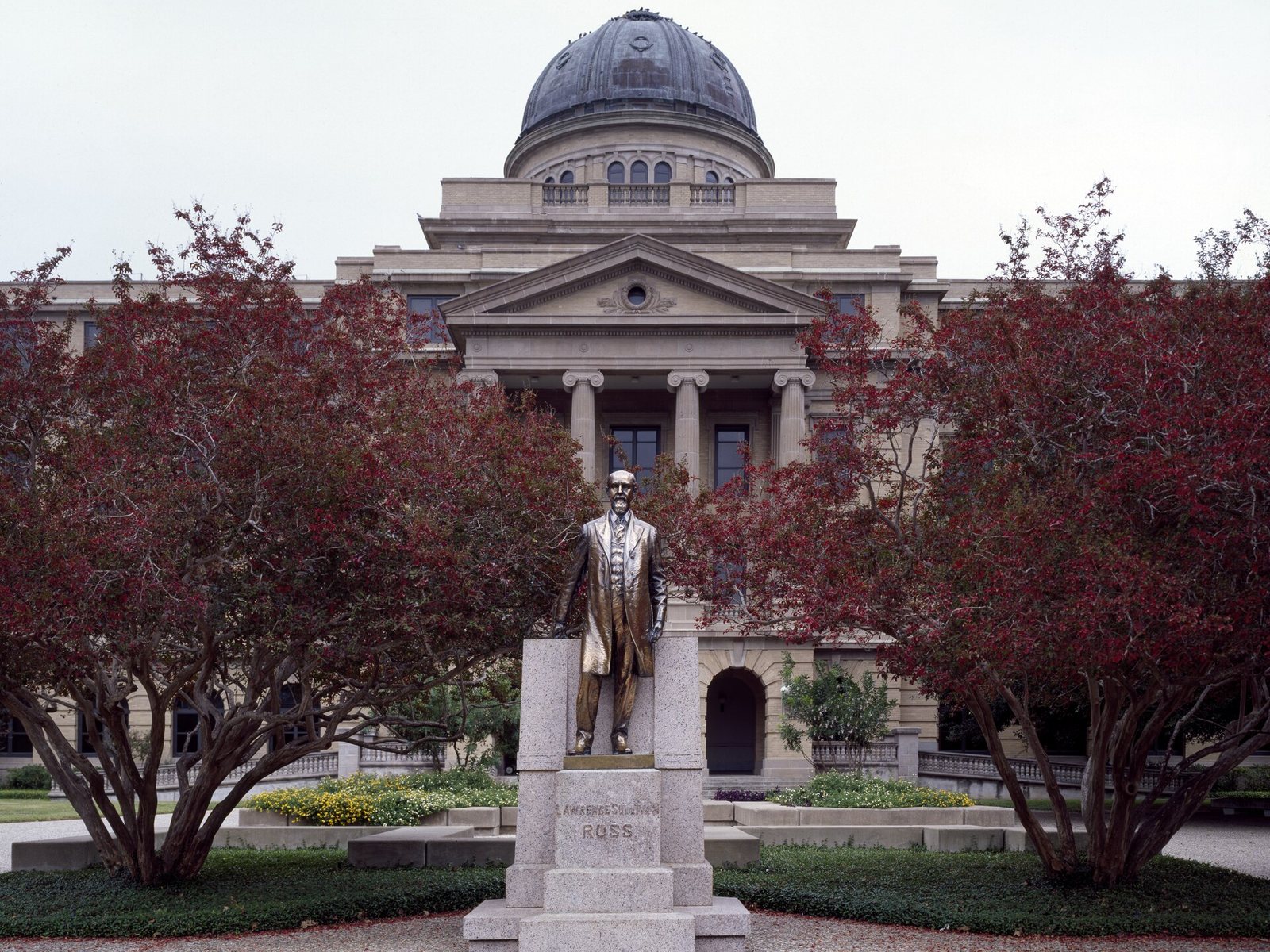 Color photograph of the bronze statue of Lawrence Sullivan Ross at Texas A&M University: a bearded, balding man in a long frock coat stands bareheaded on a gray granite pedestal carved LAWRENCE SULLIVAN ROSS, framed by two dark red-leaved crape myrtles, with the columned portico and weathered copper dome of the Academic Building rising behind him under a pale overcast sky.