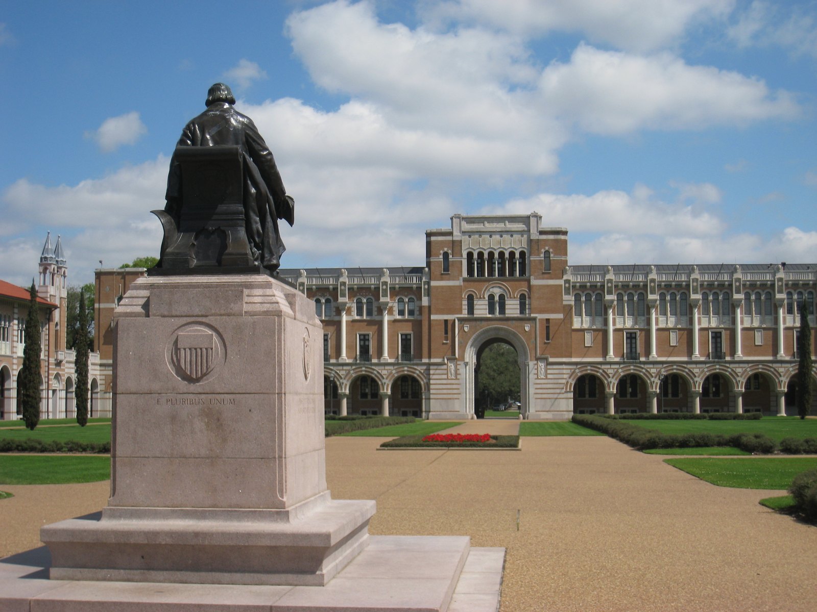Color photograph of the bronze Founder's Memorial statue of William Marsh Rice at Rice University, seen from behind: the seated figure in a long coat rests on a tall pink granite pedestal carved with a shield and the words E PLURIBUS UNUM, and looks across a broad gravel court and clipped lawns toward the arched Sallyport and long arcaded front of Lovett Hall under a blue sky with white clouds.