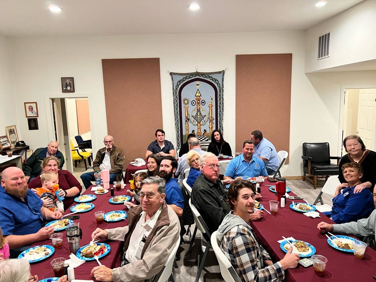 Multi-generational lodge dinner — members, wives, children, and elders around a long table beneath a Masonic tapestry.